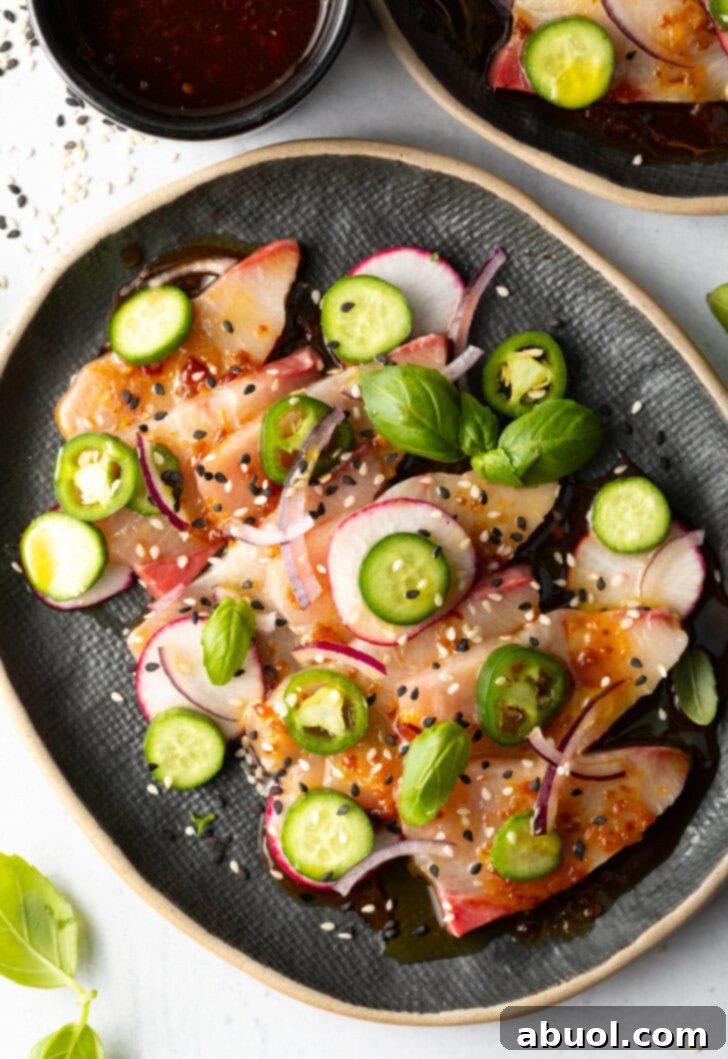 Top down view of black oval plate loaded with thin slices of yellowtail crudo topped with seasonings, thin slides of radishes, jalapenos, red onion, and a sprinkle of sesame seeds.