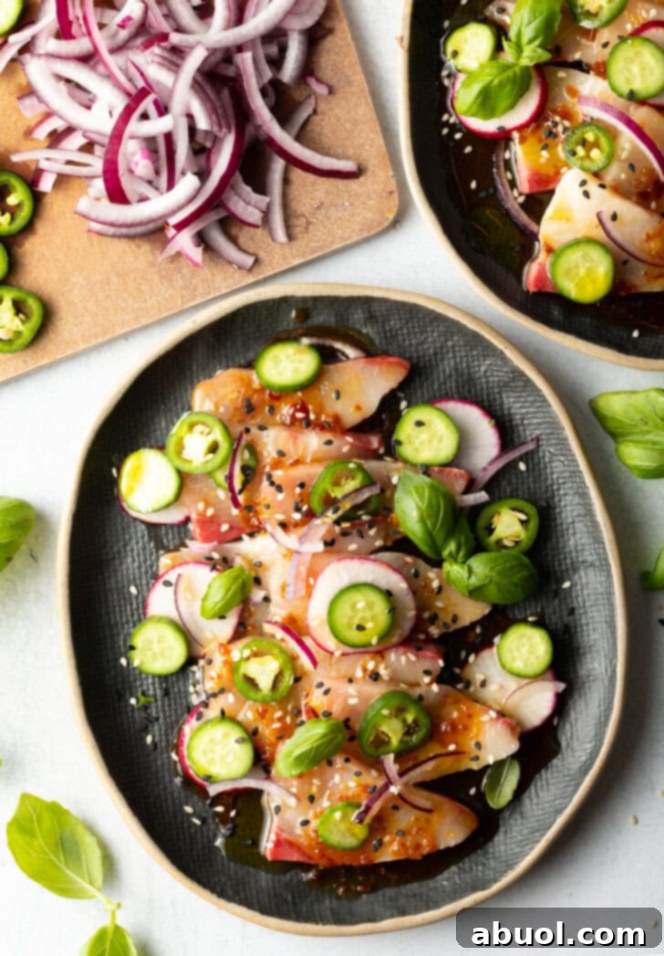 Top down view of black oval plate loaded with thin slices of yellowtail crudo topped with seasonings, thin slides of radishes, jalapenos, red onion, and a sprinkle of sesame seeds.