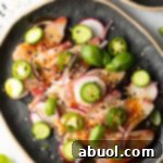 Top down view of black oval plate loaded with thin slices of yellowtail crudo topped with seasonings, thin slides of radishes, jalapenos, red onion, and a sprinkle of sesame seeds.