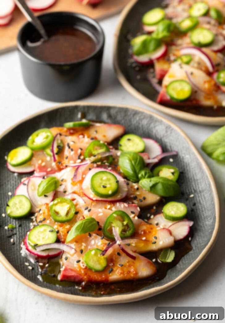 Black oval plate loaded with thin slices of hamachi crudo topped with seasonings, thin slides of radishes, jalapenos, red onion, and a sprinkle of sesame seeds.