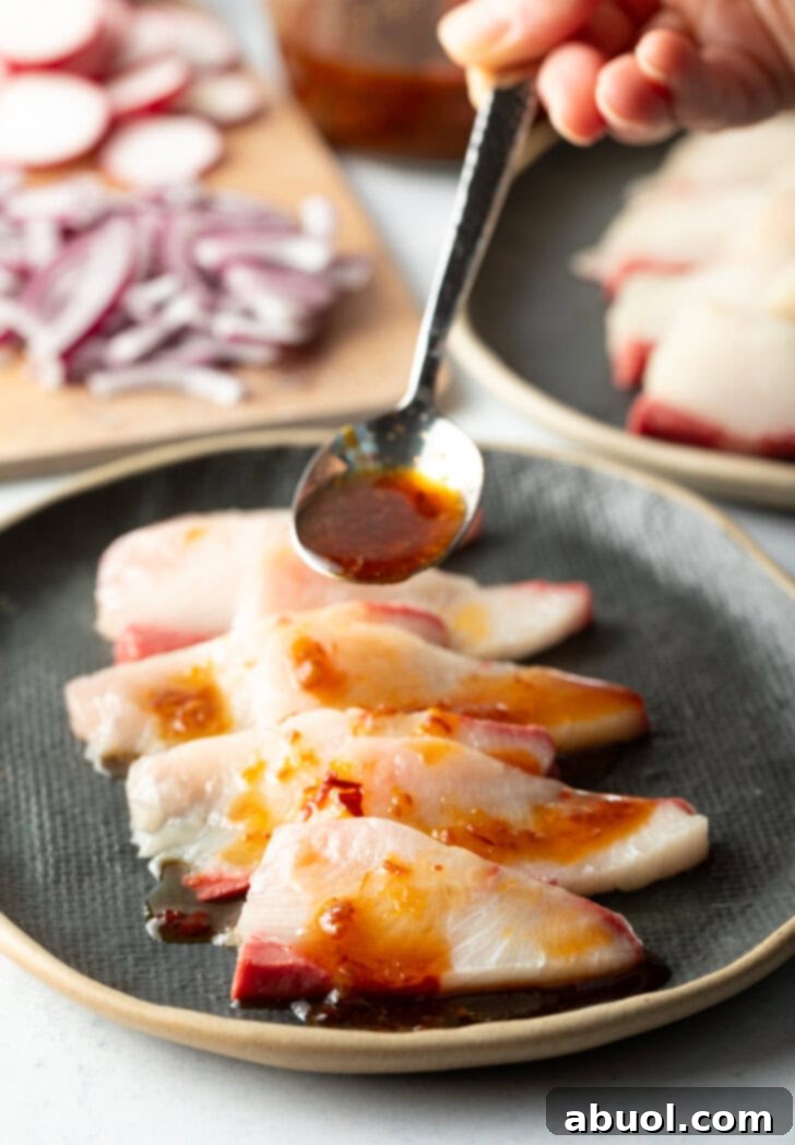 4 pieces of hamachi on a black oval plate. Spoon is adding chili soy sauce on top.