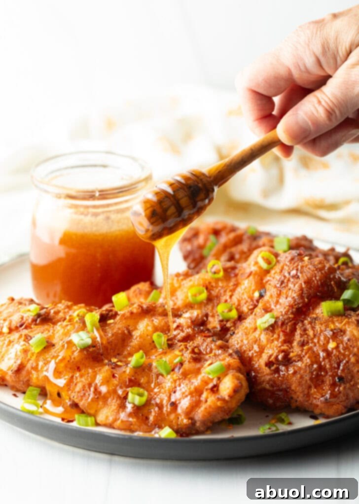 Sweet Heat Honey Blend 11 Plate of fried chicken tenders with small glass jar of hot honey to the side, A hand with a wooden honey spoon is drizzling honey over the breaded fried tenders.