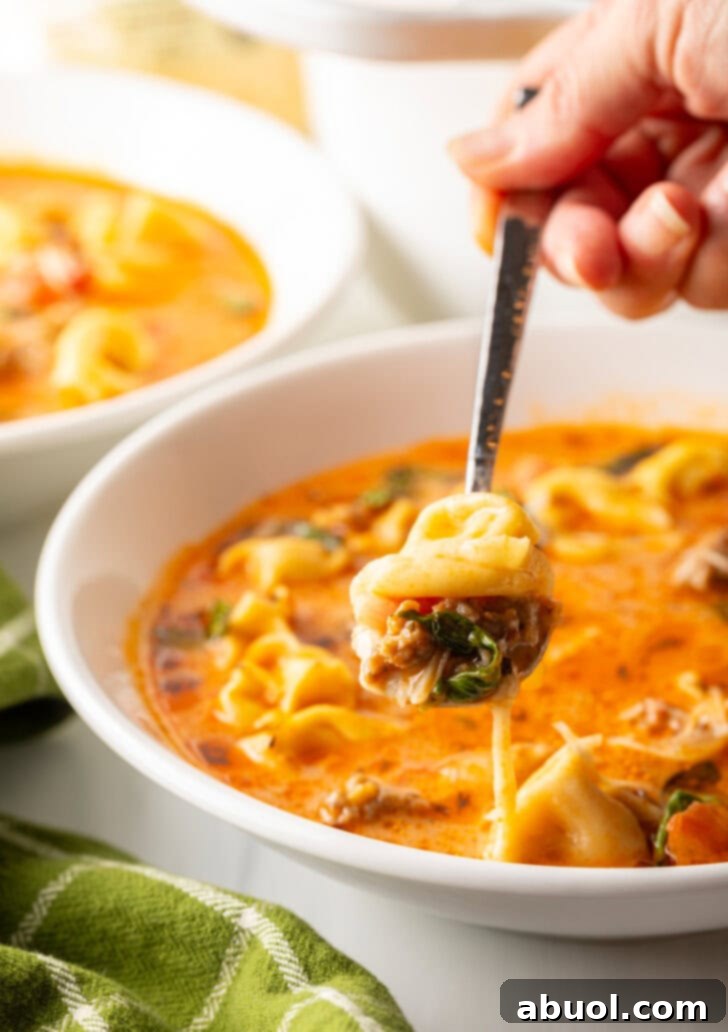 A hand holding a metal spoon, scooping a delicious bite of tortellini and fresh basil from a bowl of soup, showing its rich ingredients.