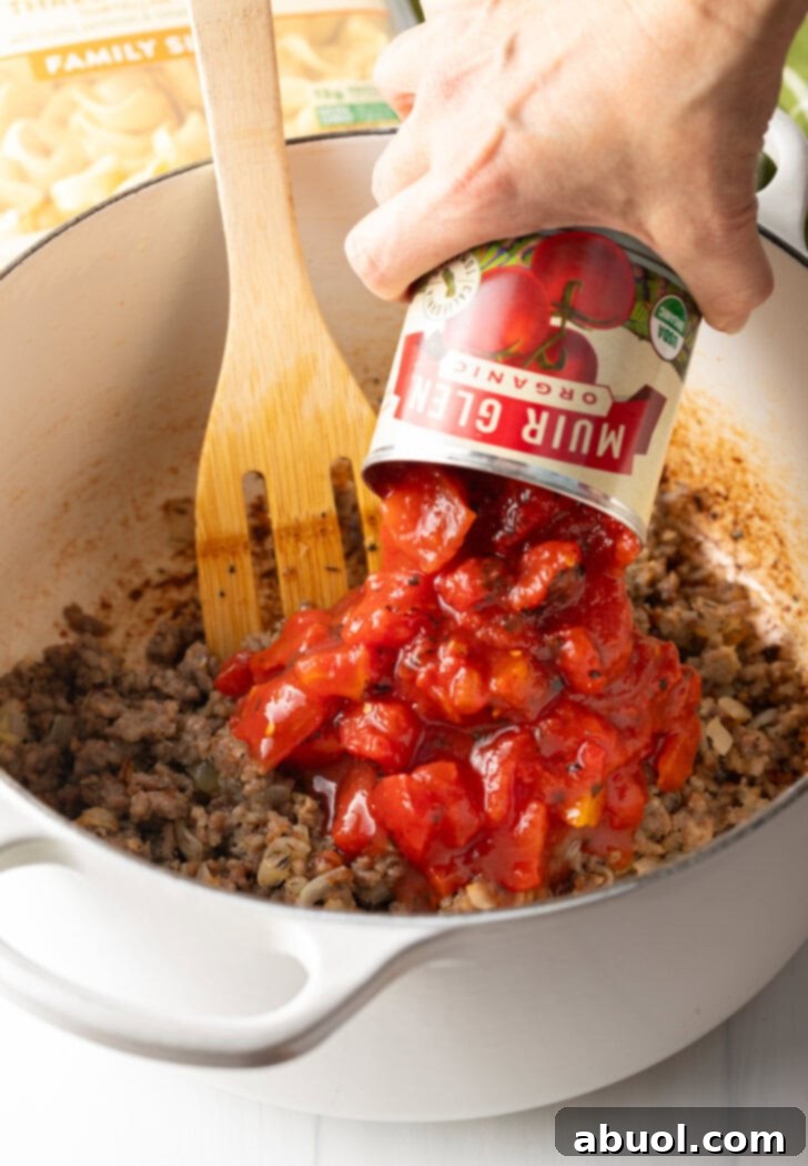 A hand pouring diced tomatoes from a can directly into the soup pot containing browned sausage, with a wooden spatula present.