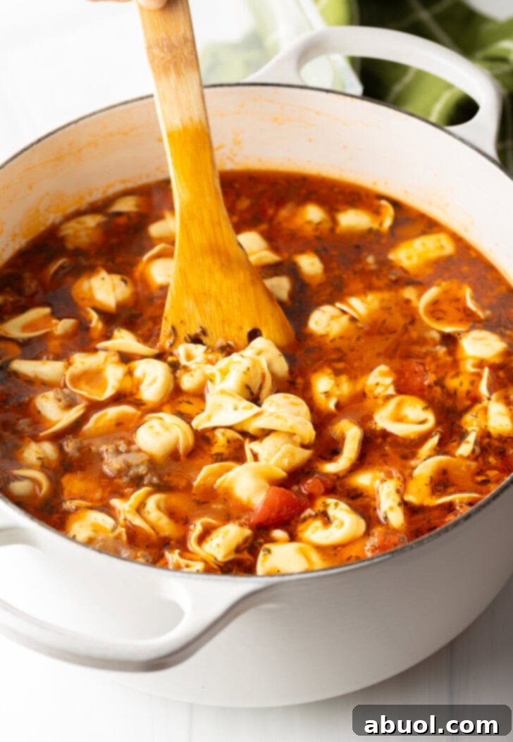 A brown spatula stirring the simmering soup in a large white pot, showing the tortellini beginning to cook.