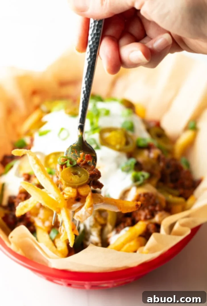 Basket with parchment paper, filled with crispy fries topped with chili and melted cheddar cheese, sour cream, chopped green onions, and jalapeno slices. A hand with forkful of fries is showing to camera.