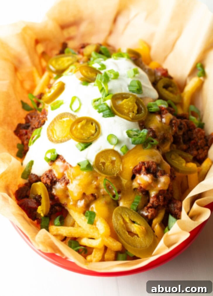 Basket with parchment paper, filled with crispy fries topped with chili and melted cheddar cheese, sour cream, chopped green onions, and jalapeno slices.