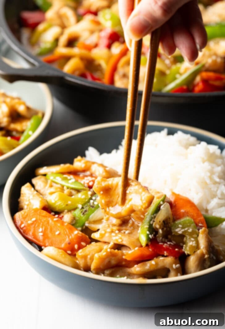 Hand with chopsticks grabbing a piece of chicken from a bowl of chop suey with white rice.