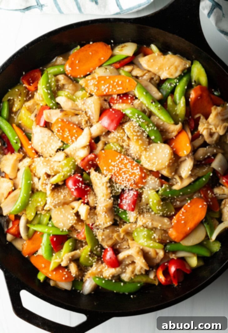 Cast iron skillet with cooked pieces of chicken, snap peas, red bell pepper, carrot slices, chopped onion, and minced garlic.