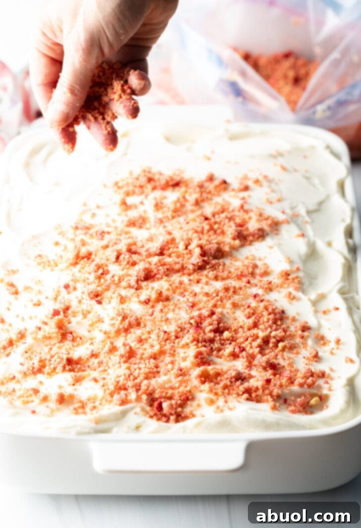 Berry Crumble Bliss Cake 12 A hand gracefully sprinkling the vibrant, crushed strawberry and cookie mixture evenly over the smooth white frosted cake, creating the signature crunch layer.