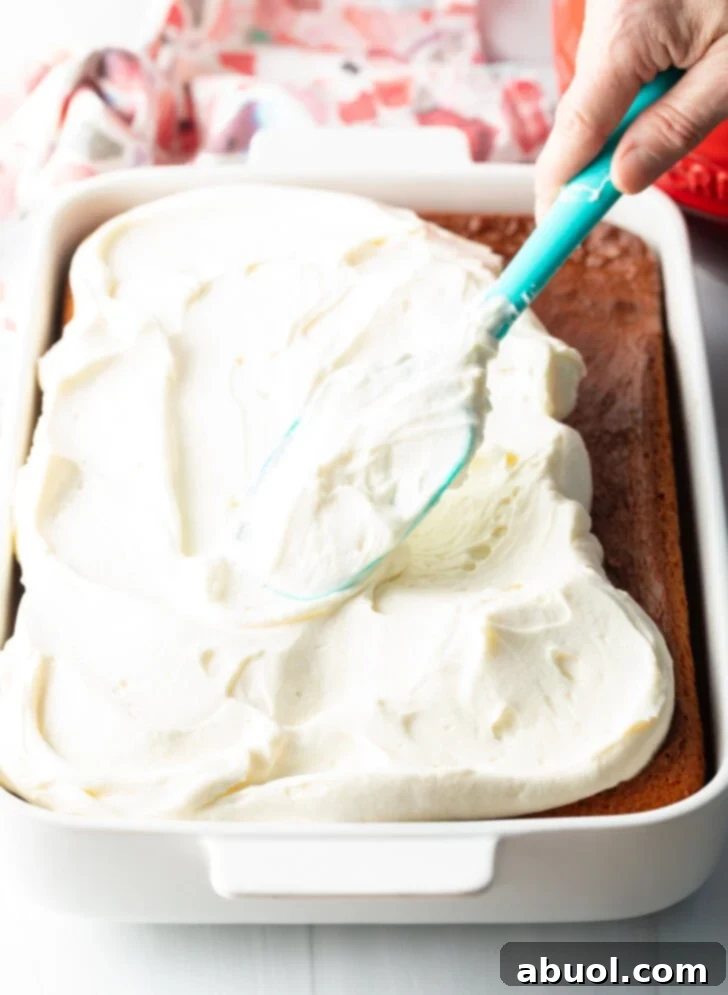 Berry Crumble Bliss Cake 9 A hand expertly spreading creamy vanilla frosting across the surface of a rectangular baked cake with a blue spatula, creating an even, smooth layer.