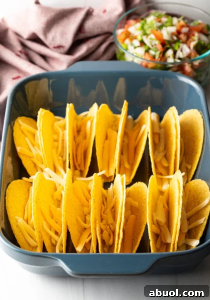 Two rows of a dozen yellow corn hard shell tacos, standing upright in a large baking dish. Shredded cheese is in the tacos.