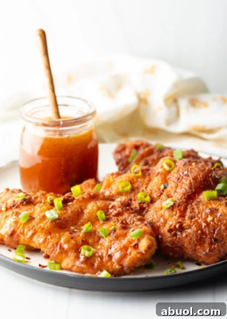 Blazing Honey Chicken 2 Three pieces of golden-brown fried hot honey chicken on a plate, garnished with vibrant sliced green onions. In the soft-focus background, a jar of glistening honey with a wooden dipper hints at the sauce's sweet origins.