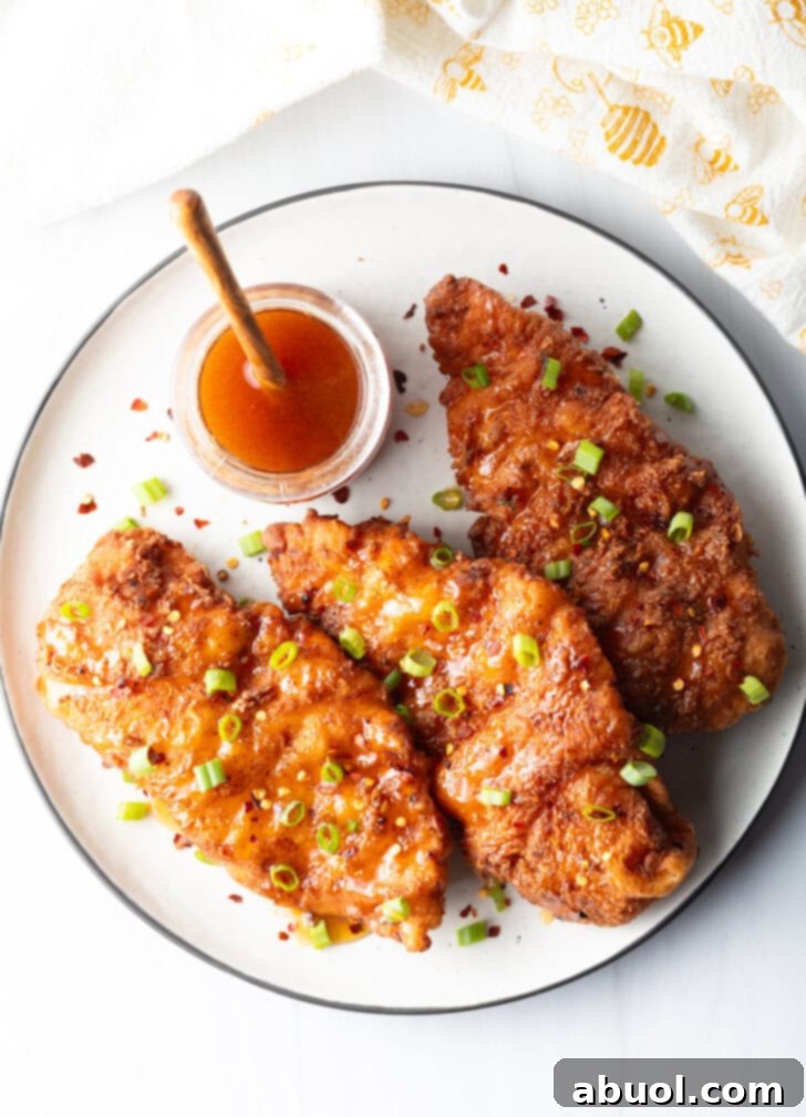Blazing Honey Chicken 11 An overhead view of three perfectly fried hot honey chicken pieces on a plate, garnished with sliced green onions. A jar of honey with a wooden spoon rests in the background, suggesting the sweet element of the dish.