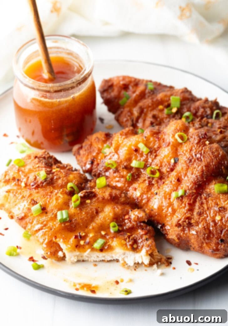 Blazing Honey Chicken 12 Three pieces of fried hot honey chicken on a plate, garnished with sliced green onions. A jar of honey is in the background, and the front piece of chicken has been neatly cut in half to reveal its juicy interior.