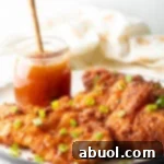 Three pieces of fried hot honey chicken on a plate. Chicken is sprinkled with sliced green onions, a jar of honey with a wood spoon in it is in the background.
