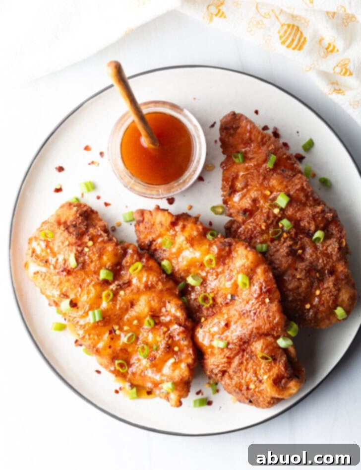 Blazing Honey Chicken 3 An overhead view of three delectable pieces of fried hot honey chicken, elegantly arranged on a plate and brightened with a sprinkle of sliced green onions. A rustic jar of honey with a wooden spoon sits invitingly in the background, symbolizing the key ingredient.