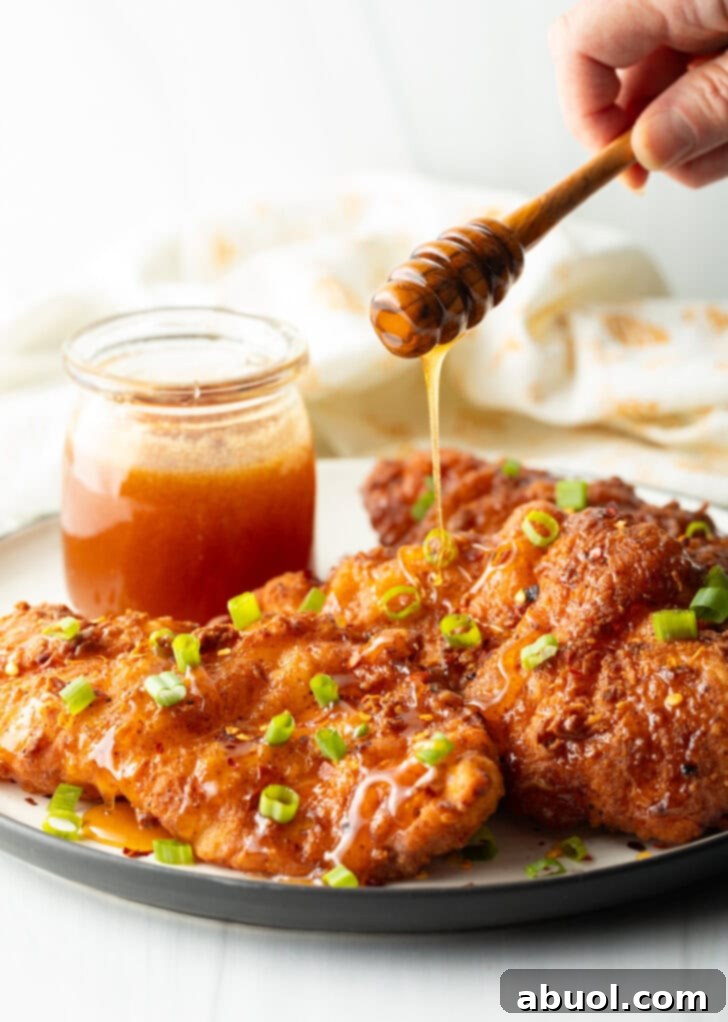 Blazing Honey Chicken 4 A close-up shot of three pieces of golden-fried hot honey chicken on a plate, adorned with sliced green onions. A hand is artfully drizzling hot honey over one piece, creating glistening streaks of sweet and spicy goodness.