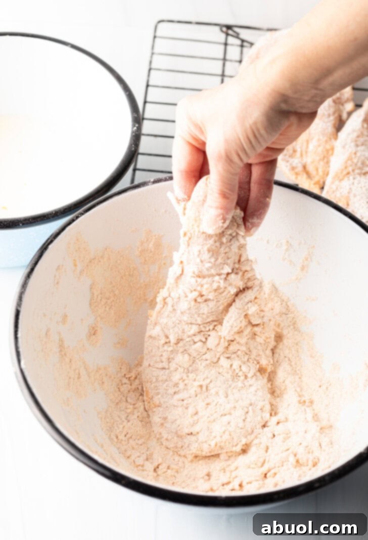 Blazing Honey Chicken 6 A hand is gently dredging a thin chicken breast cutlet in a bowl of seasoned flour, ensuring an even coating for the first step of breading.