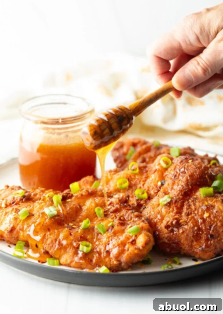 Blazing Honey Chicken 10 Three pieces of perfectly fried hot honey chicken on a plate, beautifully garnished with sliced green onions. In the background, a jar of honey sits. A hand is actively drizzling hot honey over the chicken, enhancing its glossy appeal.