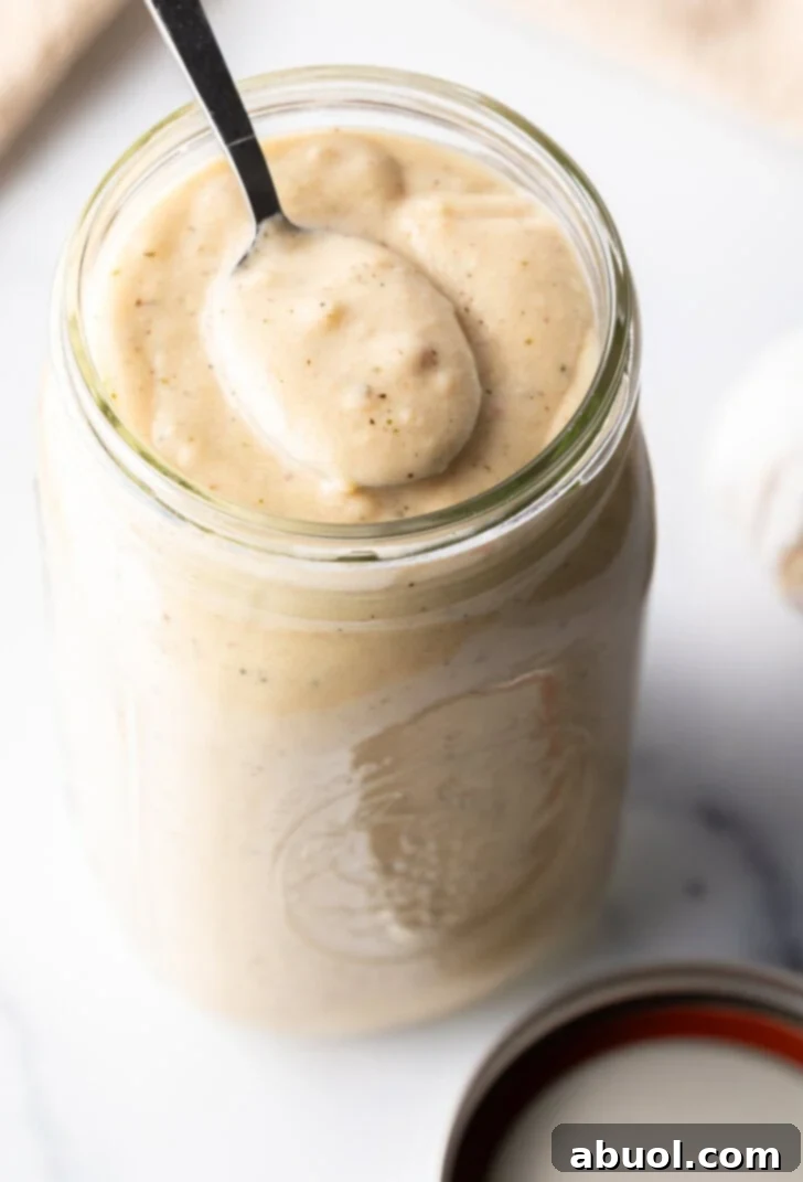 Large glass mason jar filled with homemade garlic and parmesan sauce. A metal spoon is showing a scoop of sauce to camera.