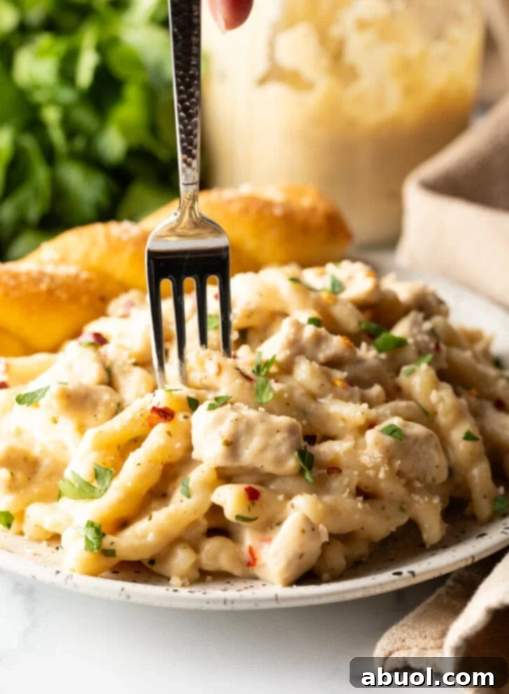 White plate of cheesy garlic parmesan chicken pasta with a breadstick twist on the side of the plate. A fork is taking a bite of pasta from the plate.