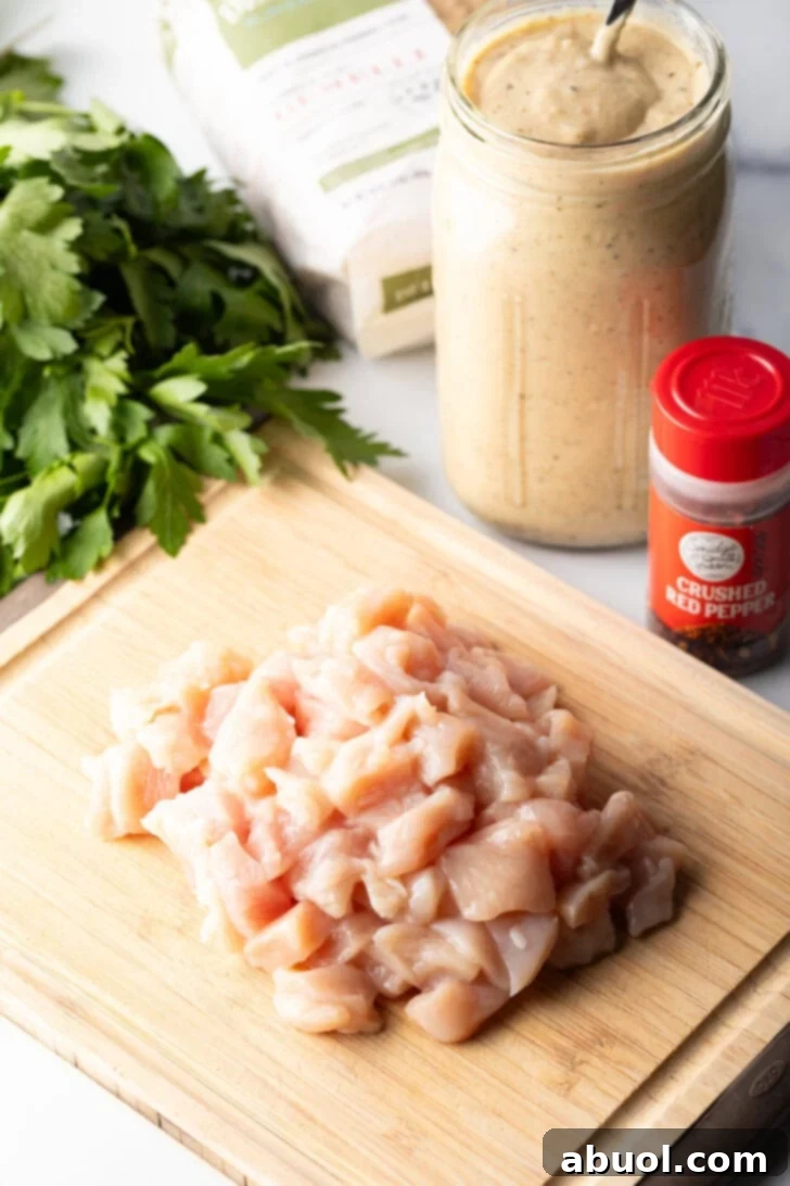 Mound of diced chicken breast pieces on a wood cutting board. In the background is a jar of garlic parmesan sauce, a bottle of seasoning, and fresh parsley leaves.