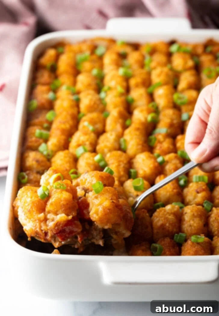 Full baked casserole with rows of tater tots on top. A hand with a metal spoon is scooping a serving.