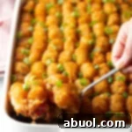 Full baked casserole with rows of tater tots on top. A hand with a metal spoon is scooping a serving.