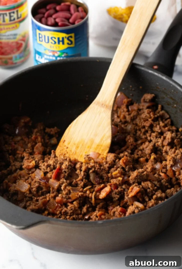 Deep black skillet with ground beef mixture, being stirred by a wooden spatula.