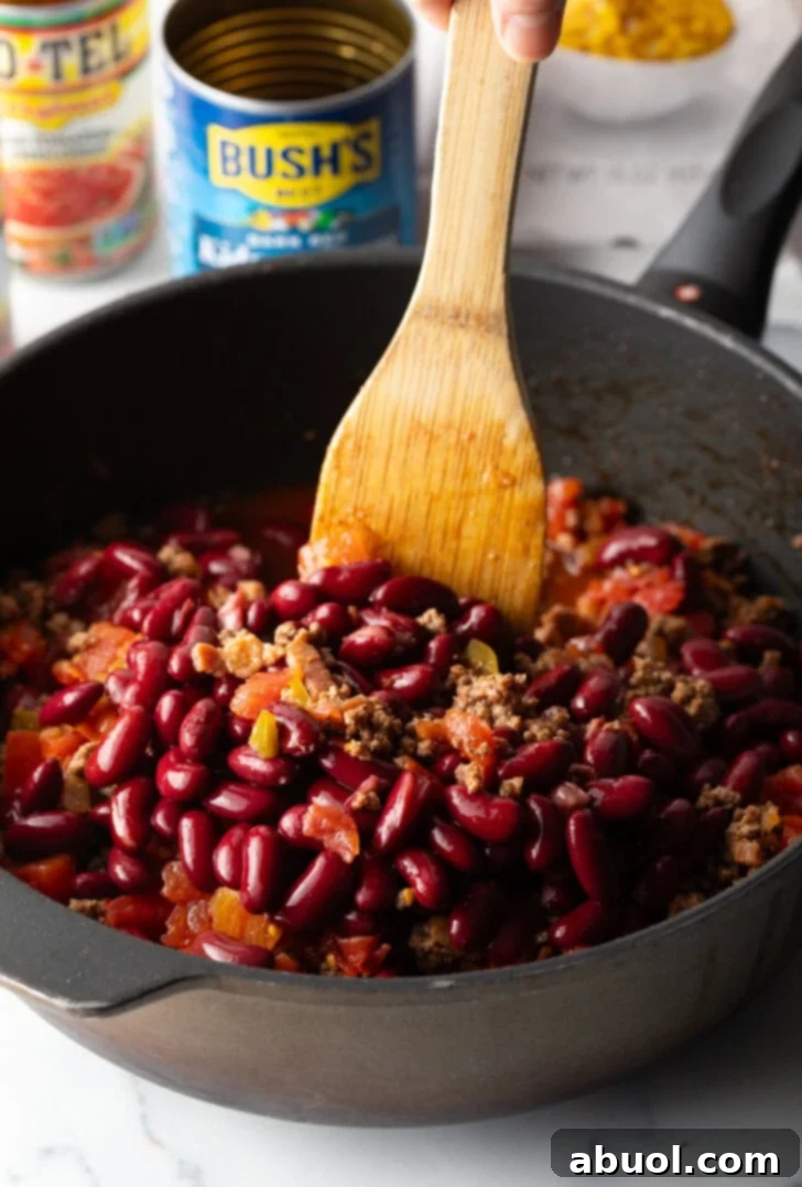 Deep black skillet with ground beef mixture and beans, being stirred by a wooden spatula.