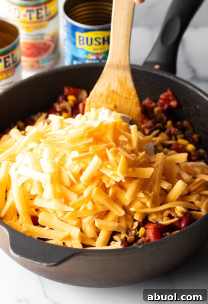 Deep black skillet with ground beef mixture and shredded cheese, being stirred by a wooden spatula.