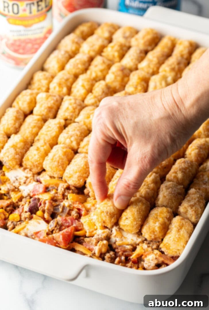 Hand adding frozen tater tots to the top of a baking dish casserole.