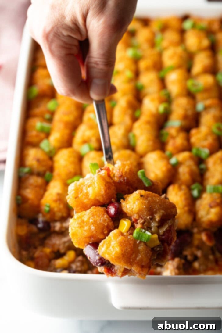 Full baked casserole with rows of tater tots on top. A hand with a metal spoon is scooping a serving.