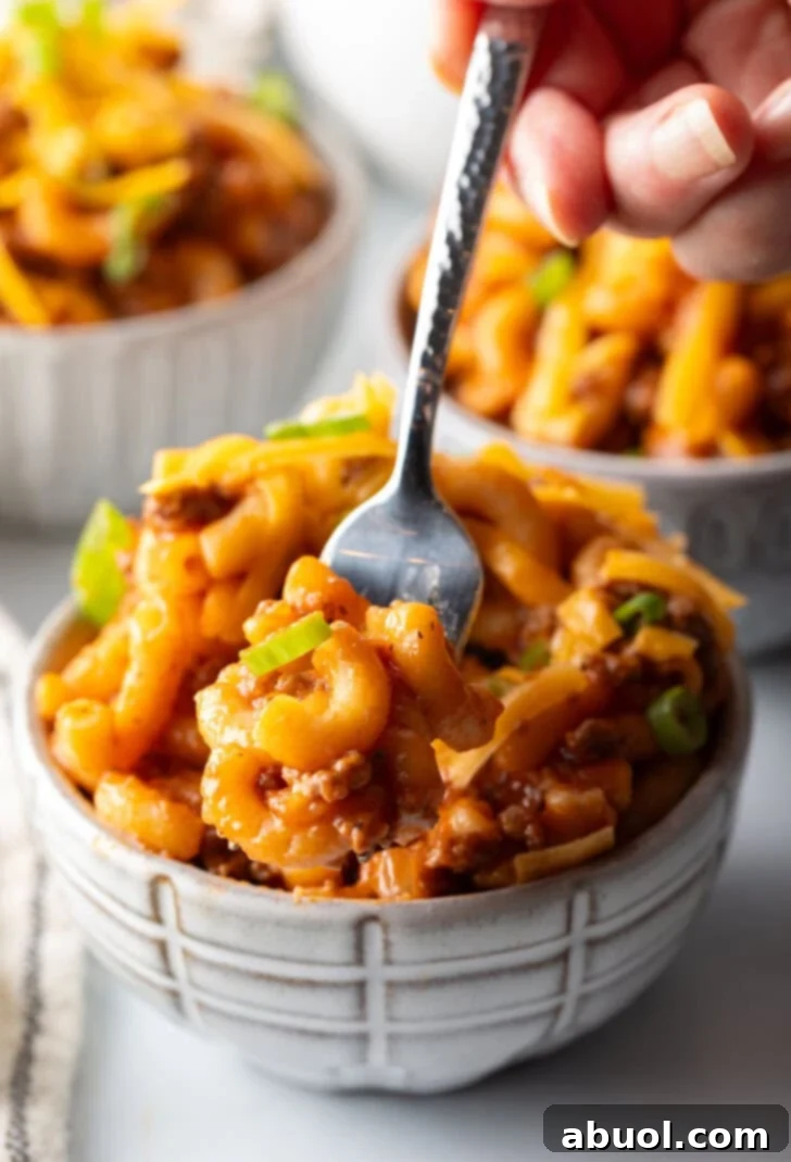 Hearty Beef and Macaroni Skillet 4 A close-up shot of a bowl of homemade beefaroni, garnished with melted shredded cheddar cheese and vibrant chopped scallions. A fork is digging in, showing the rich, saucy macaroni and ground beef below.