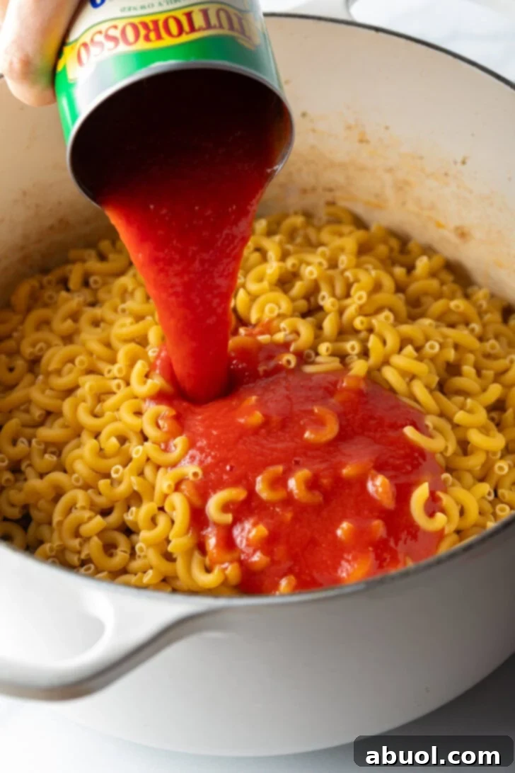 Hearty Beef and Macaroni Skillet 6 Canned tomato sauce being added to a large cooking pot containing elbow macaroni and ground beef mixture.