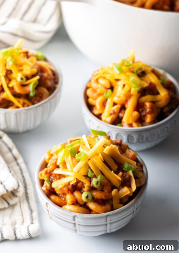 Hearty Beef and Macaroni Skillet 9 Three bowls of homemade beefaroni, generously topped with shredded cheddar cheese and vibrant chopped scallions, presented on a rustic wooden surface.