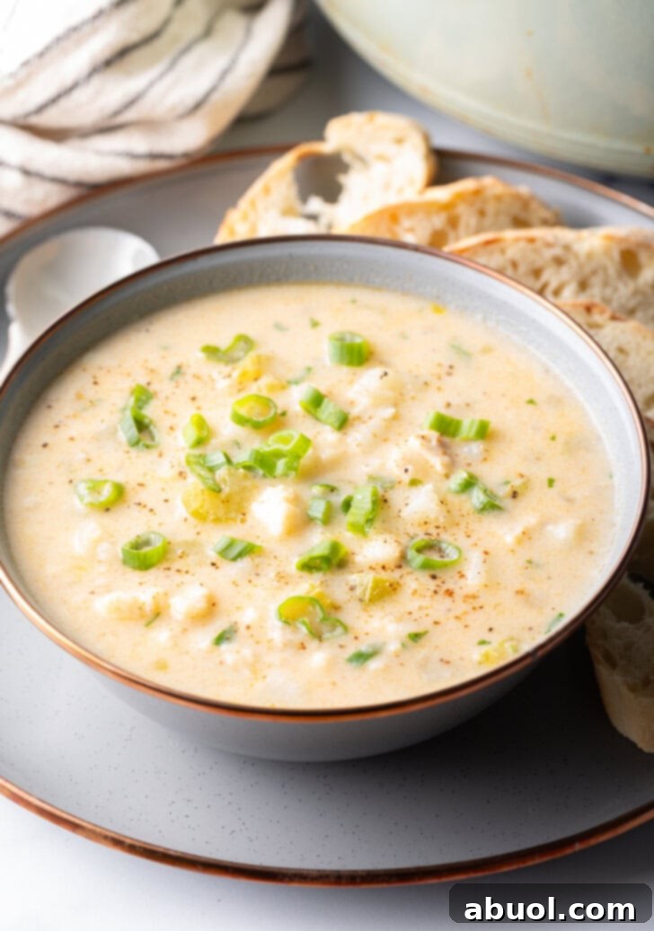 A comforting bowl of creamy fish chowder, garnished with fresh herbs, sits on a grey plate alongside slices of crusty bread and a spoon.