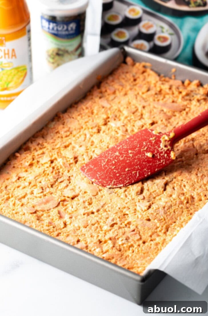 A red spatula smoothing the savory salmon and sriracha mayo mixture over the cream cheese layer in a baking pan.