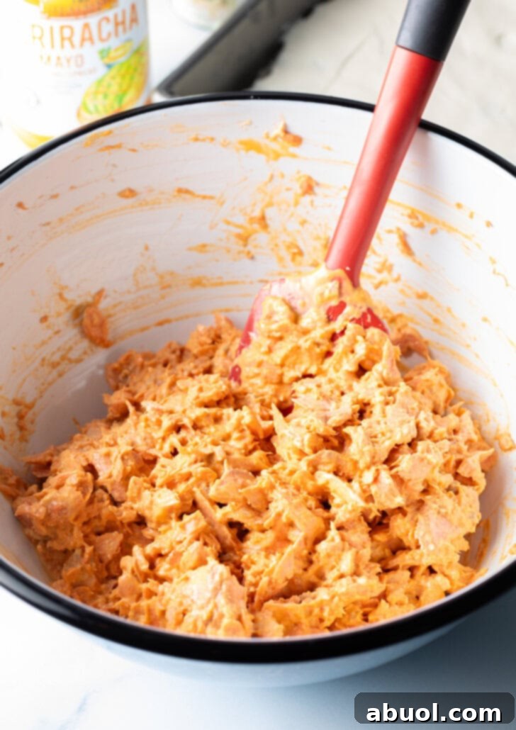 A red spatula stirring together tender flaked salmon and creamy sriracha mayonnaise in a pristine white mixing bowl.