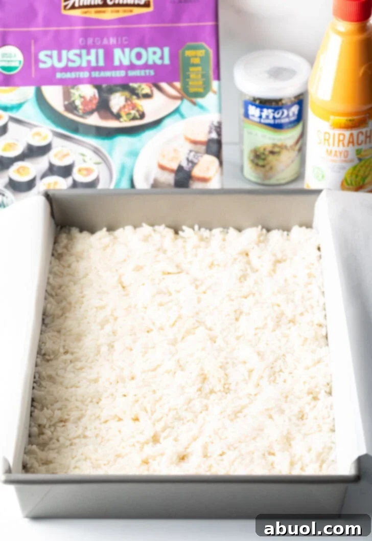 A perfectly even layer of white sushi rice firmly pressed into the bottom of a rectangular baking pan, forming the base.