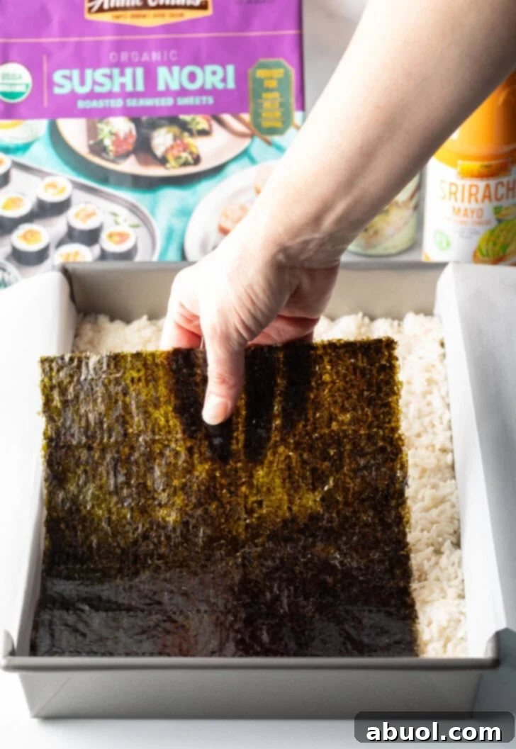 A hand carefully placing a large sheet of dark green nori seaweed over the seasoned rice layer in a baking pan.