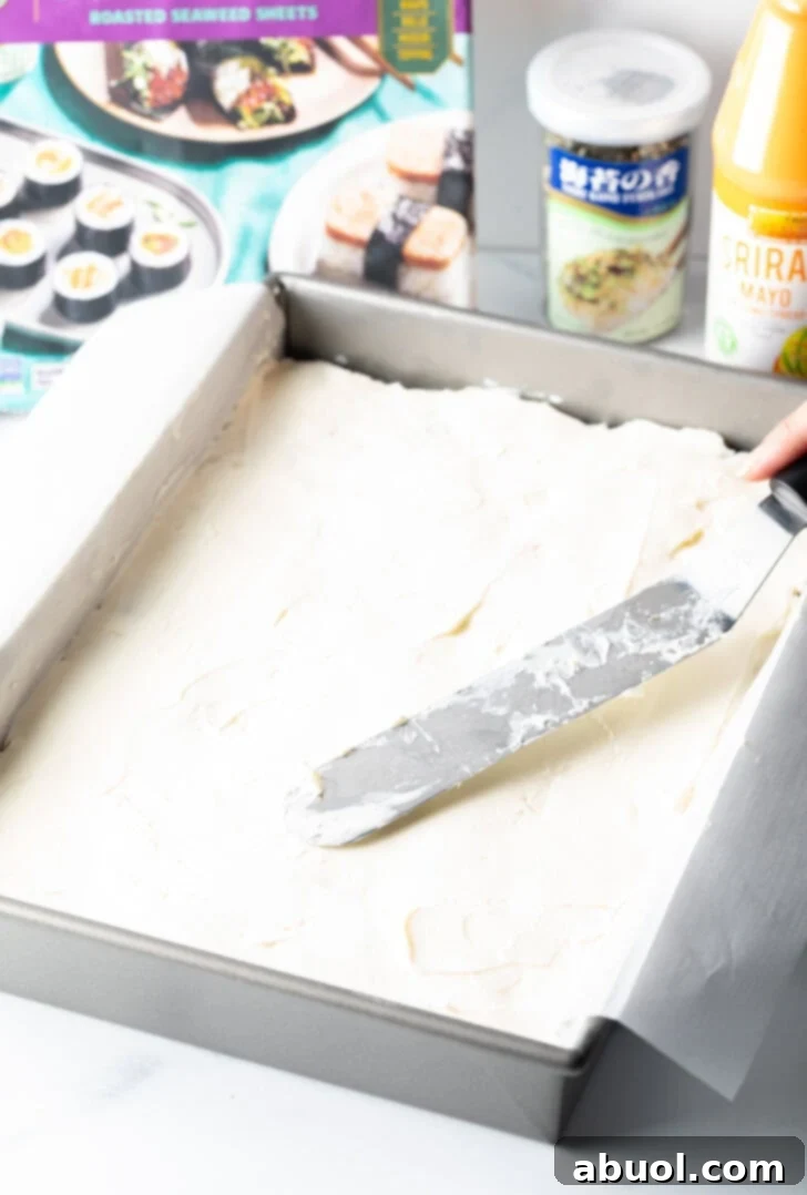 Using a flat metal spatula to smoothly spread softened cream cheese over the nori layer in a rectangular baking pan.