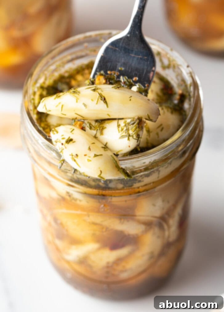 Top down open glass jar stuffed with preserved garlic and herbs. A fork is digging in to grab several cloves.