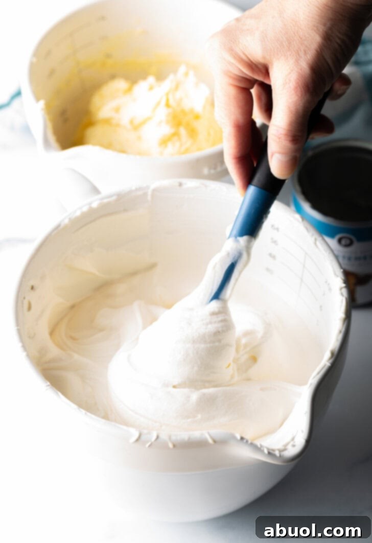 No-Churn Banana Pudding Dream 5 Hand using blue rubber spatula to blend vanilla ice cream base in a large white bowl.