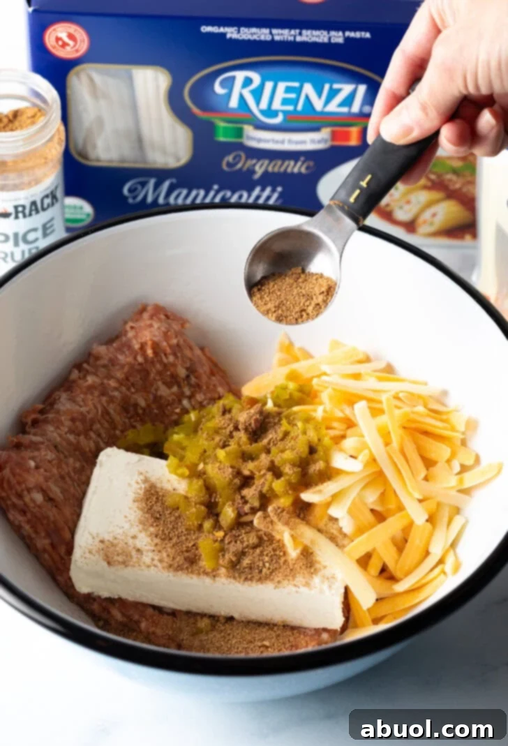 Hand adding tablespoon of seasoning to a white bowl with raw ground beef, shredded cheddar cheese, block of cream cheese, and chopped peppers.