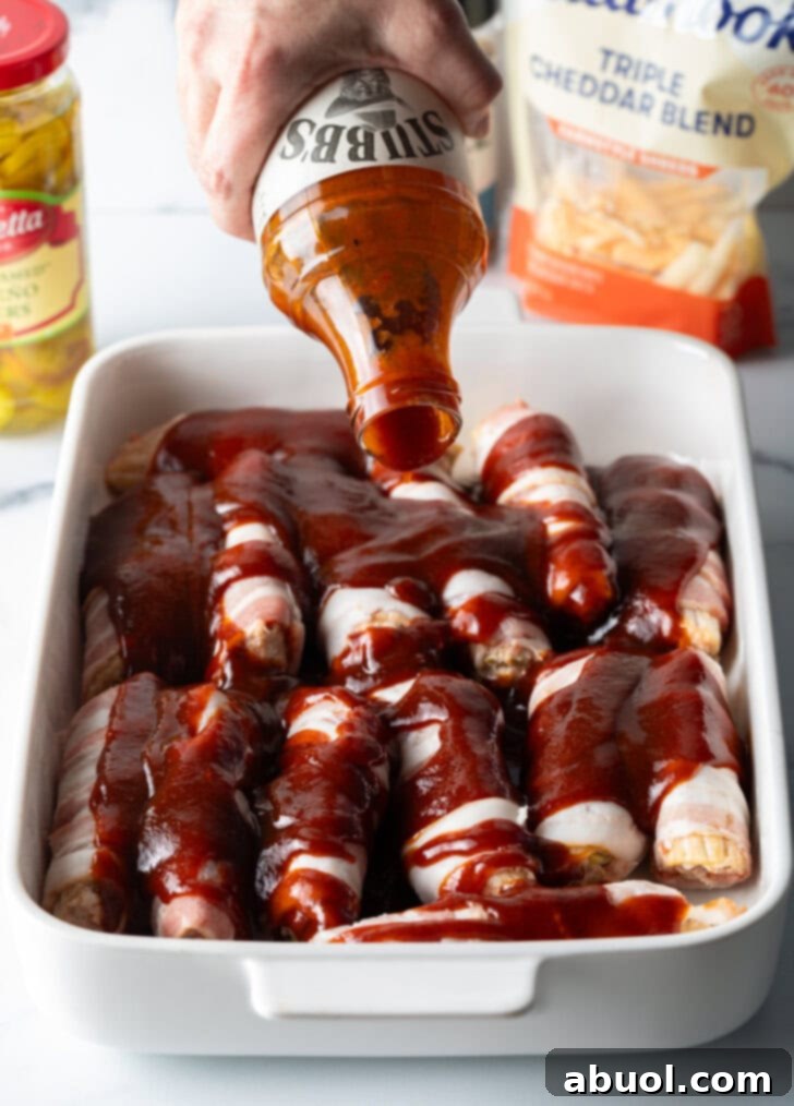 Hand pouring barbecue sauce from a bottle on top of the bacon wrapped and stuffed shells layered in a white baking dish.