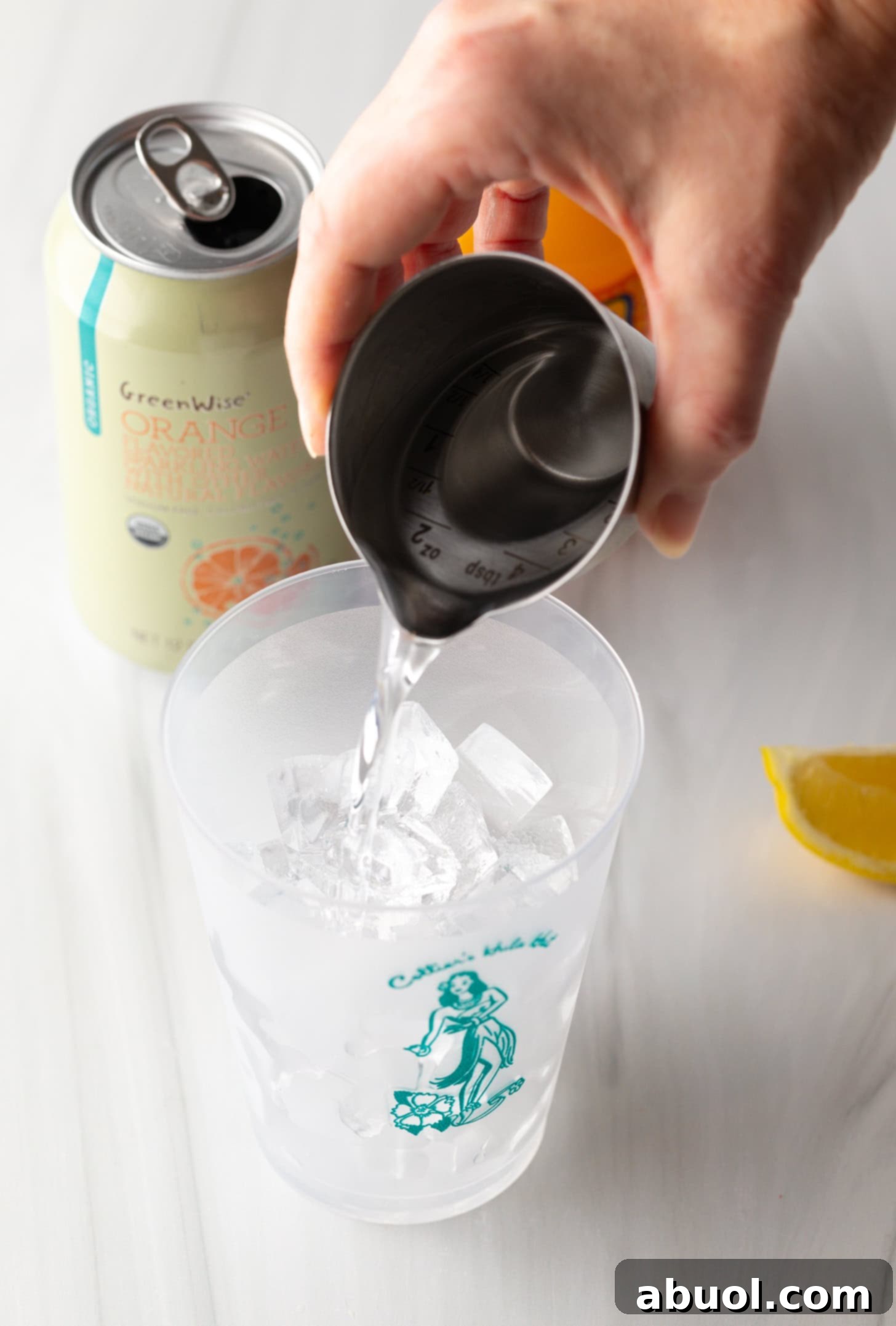 A hand pouring vodka from a metal shot glass into a clear glass filled with ice.