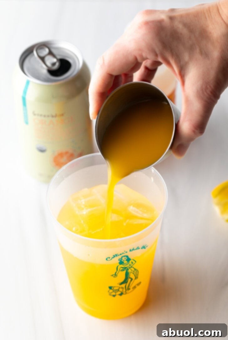 A hand adding orange juice from a metal jigger into a clear glass cup already containing ice and other liquids.