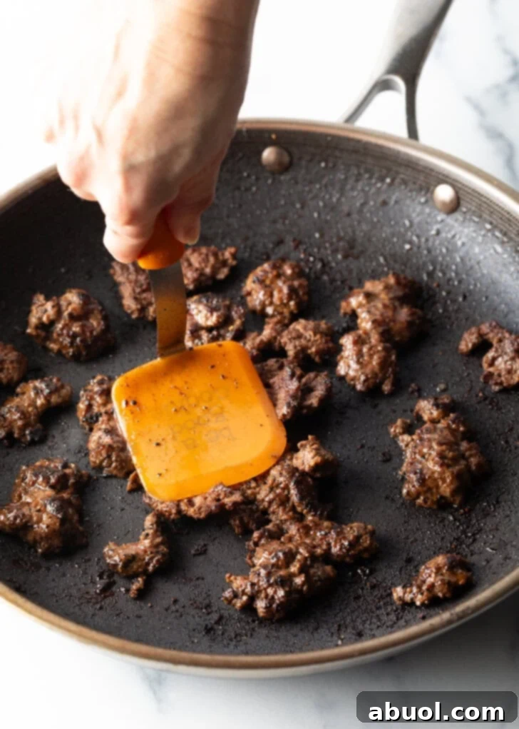 A hand expertly using a spatula to cook mini burger patties in a hot, seasoned skillet, demonstrating the browning process of the ground beef and creating crispy edges.